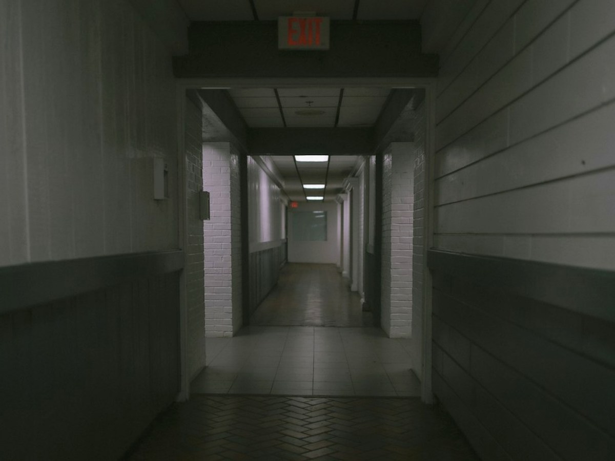 A narrow set-built corridor with a keypad panel and a heavy prop door at the end, lit from above.